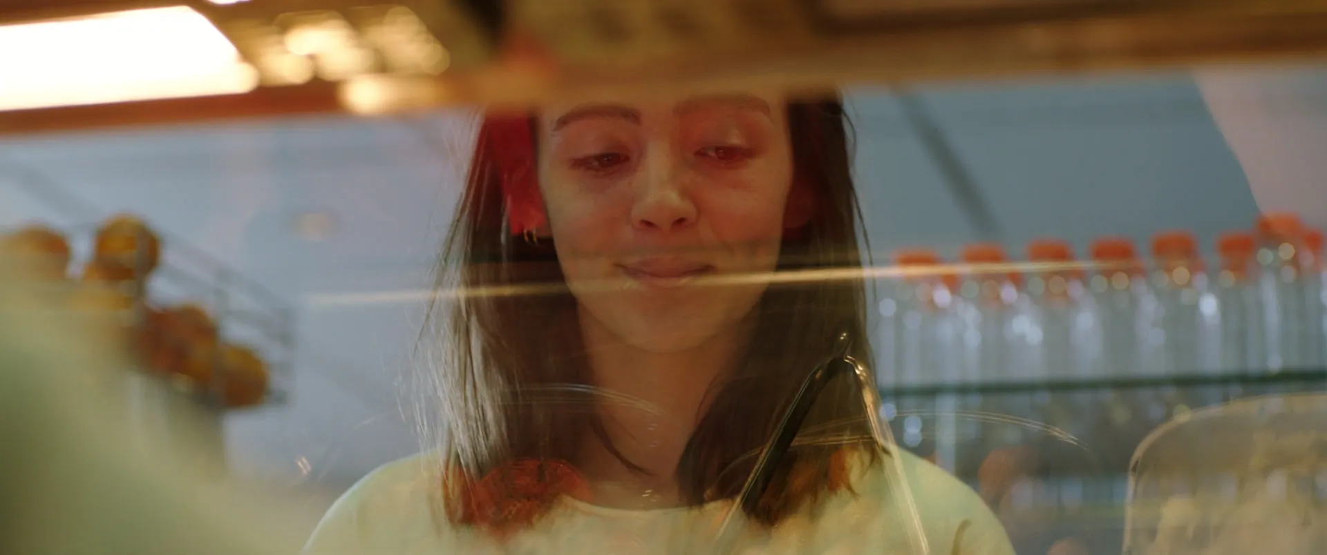 Visage d'une jeune femme vu à travers la vitre d'une station-service, brouillé par les reflets orange des rayonnages.