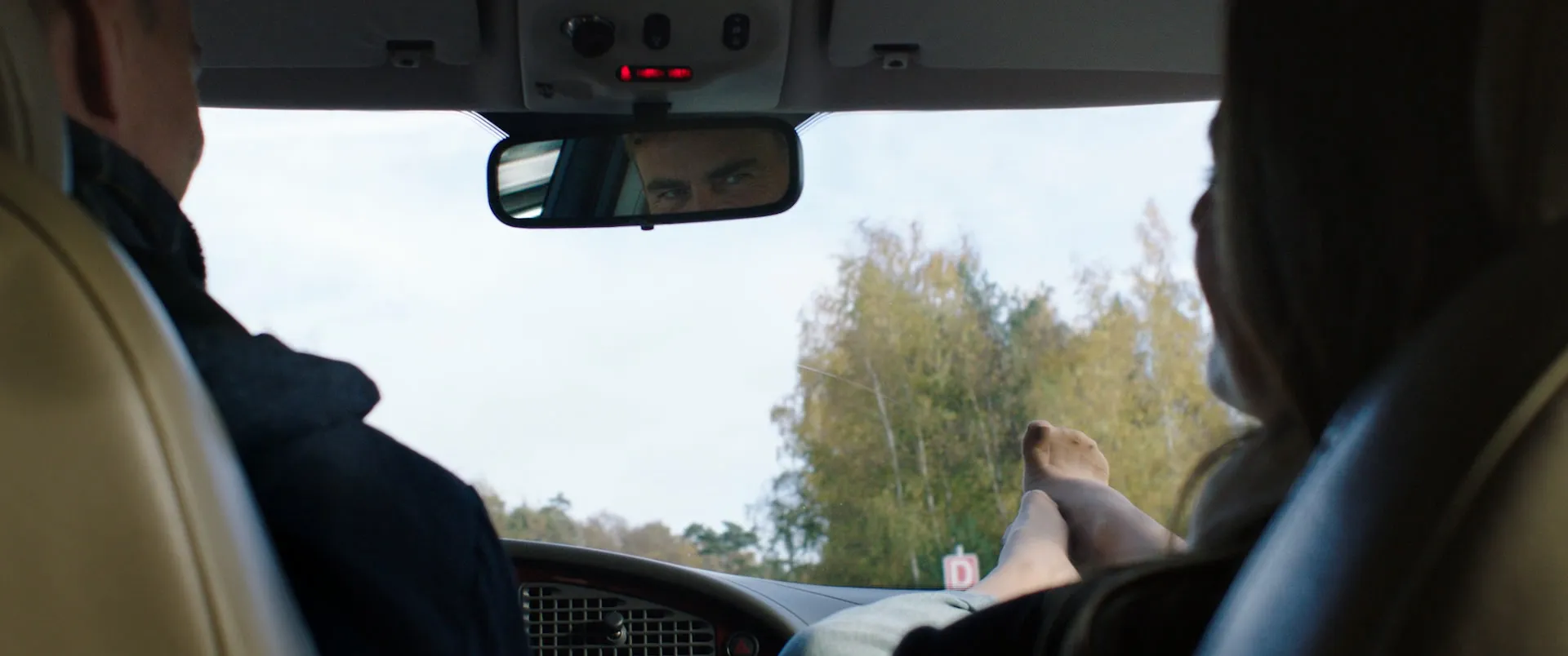 Vue depuis la banquette arrière d'une voiture : le rétroviseur découpe les yeux du conducteur, des pieds nus posés sur le tableau de bord.