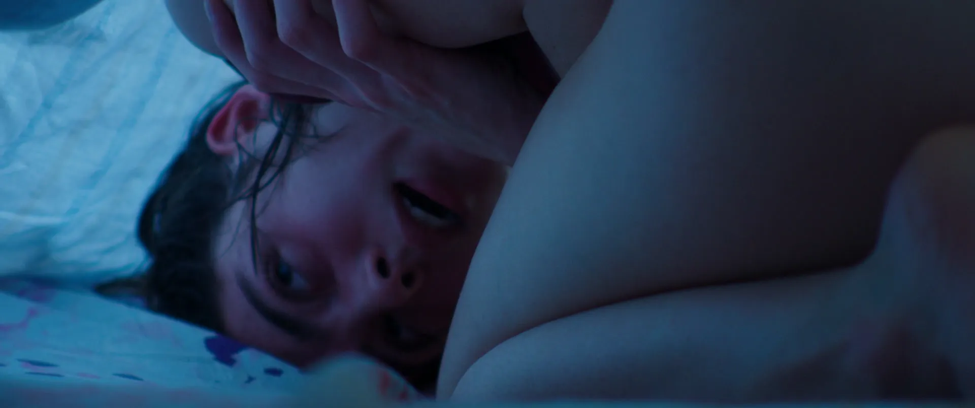 Gros plan en lumière bleue nocturne : un visage renversé contre un corps nu, les traits presque effacés par la pénombre.