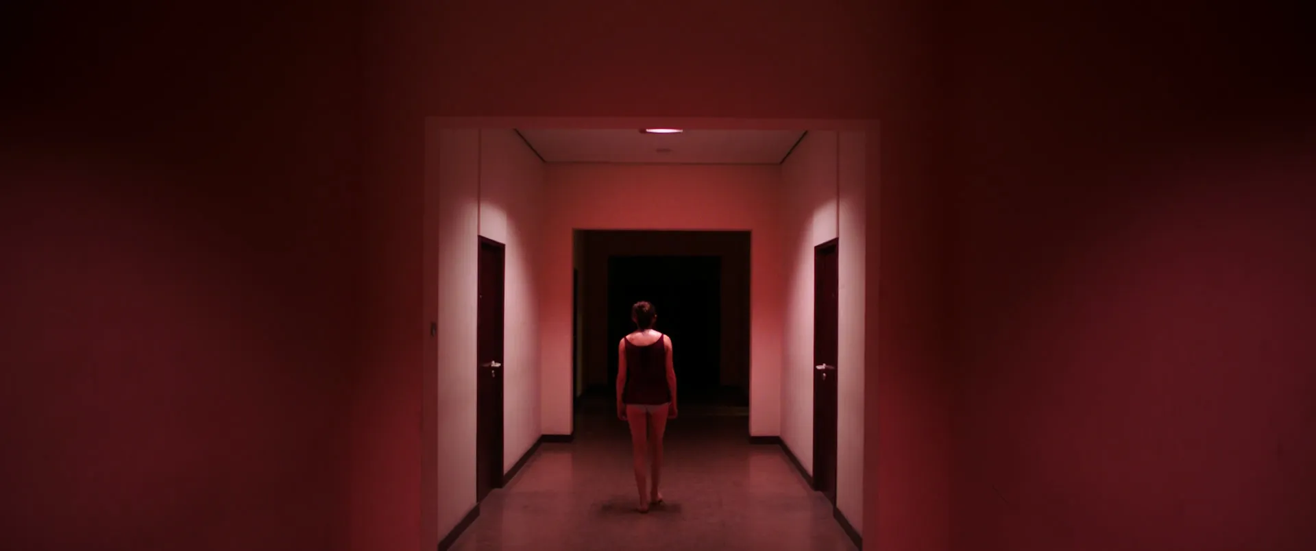 Un couloir entièrement rouge, portes fermées de part et d'autre, une silhouette de dos avançant vers l'obscurité au centre exact du cadre.