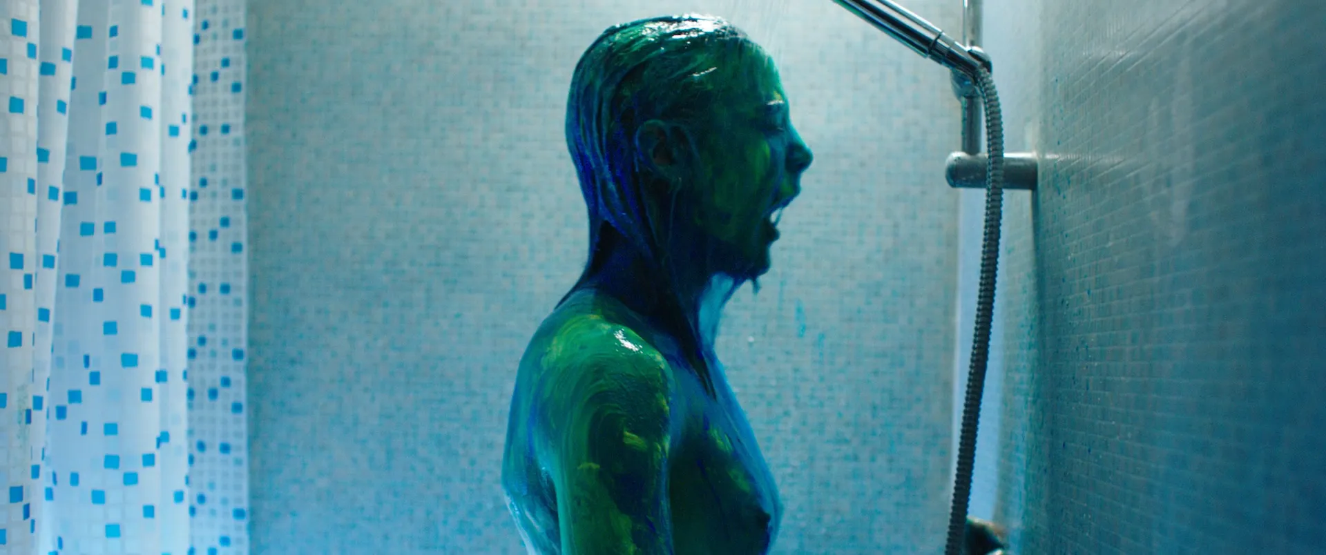 Une personne sous la douche, le corps et les cheveux couverts de peinture verte et bleue, devant un carrelage bleu clair et un rideau à damier.