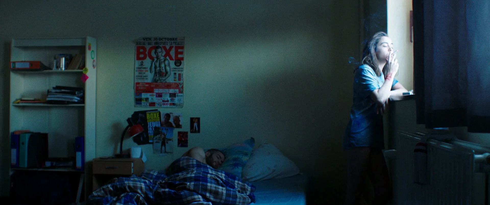 Plan large d'une chambre d'internat de nuit : une silhouette fume à la fenêtre, une autre dort sur le lit, une affiche de boxe au mur.
