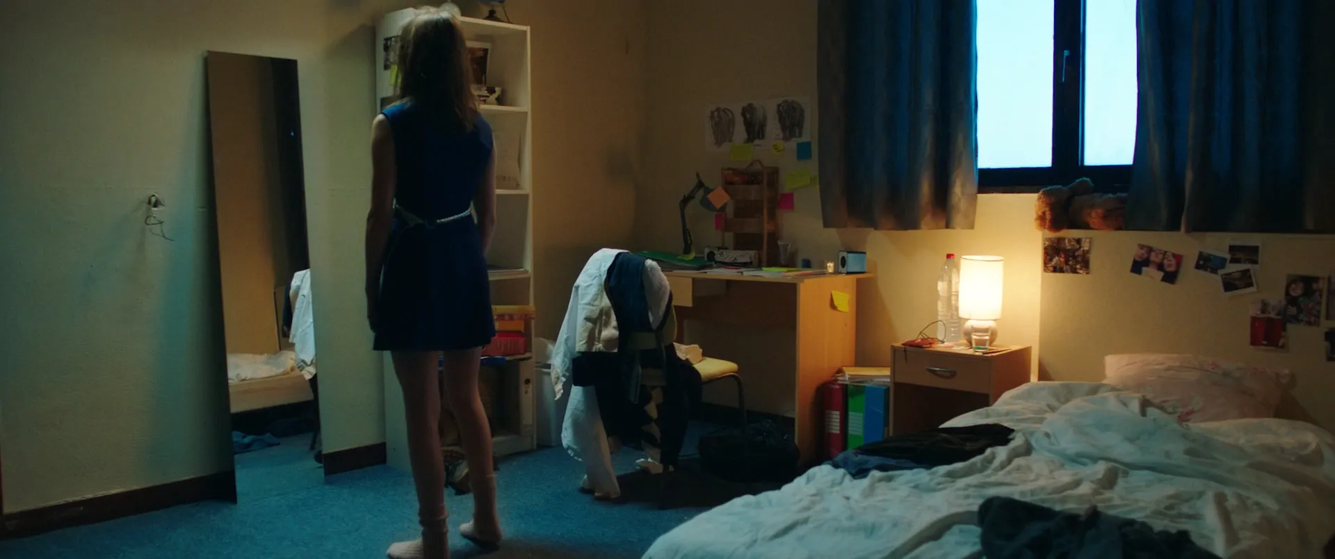 Plan large d'une chambre d'internat : une jeune femme de dos en robe bleue devant une étagère, lit défait et lampe allumée.